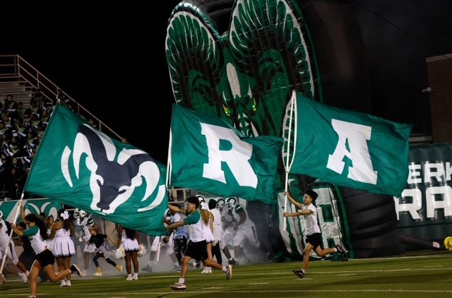 Students run across the football field carrying large green flags with the Rams logo and letters spelling “RA” as the team takes the field.