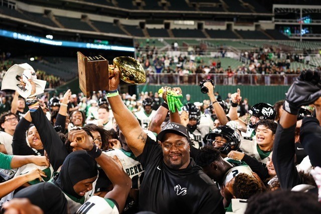 A coach raises a trophy above his head while surrounded by players celebrating on the field after a playoff win.