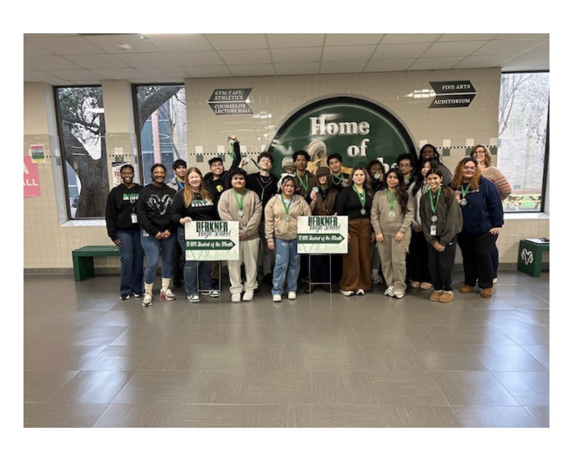 A group of diverse high school students and staff posing in a school hallway in front of a "Home of the Rams" wall graphic. Two students are holding signs that read "Berkner High School Ram Student of the Month," and many students are wearing commemorative medals on green ribbons.