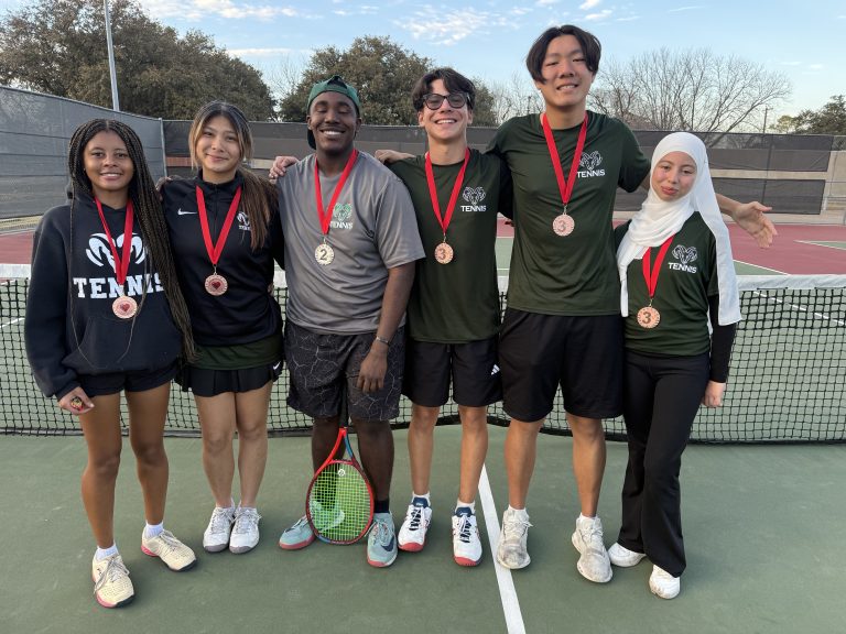 A group photo of six smiling varsity tennis players standing on an outdoor tennis court in front of the net. All six athletes are wearing medals with red ribbons around their necks. They are dressed in team gear, including dark green "TENNIS" shirts with a ram logo and black athletic wear. One player in the center holds a tennis racket.