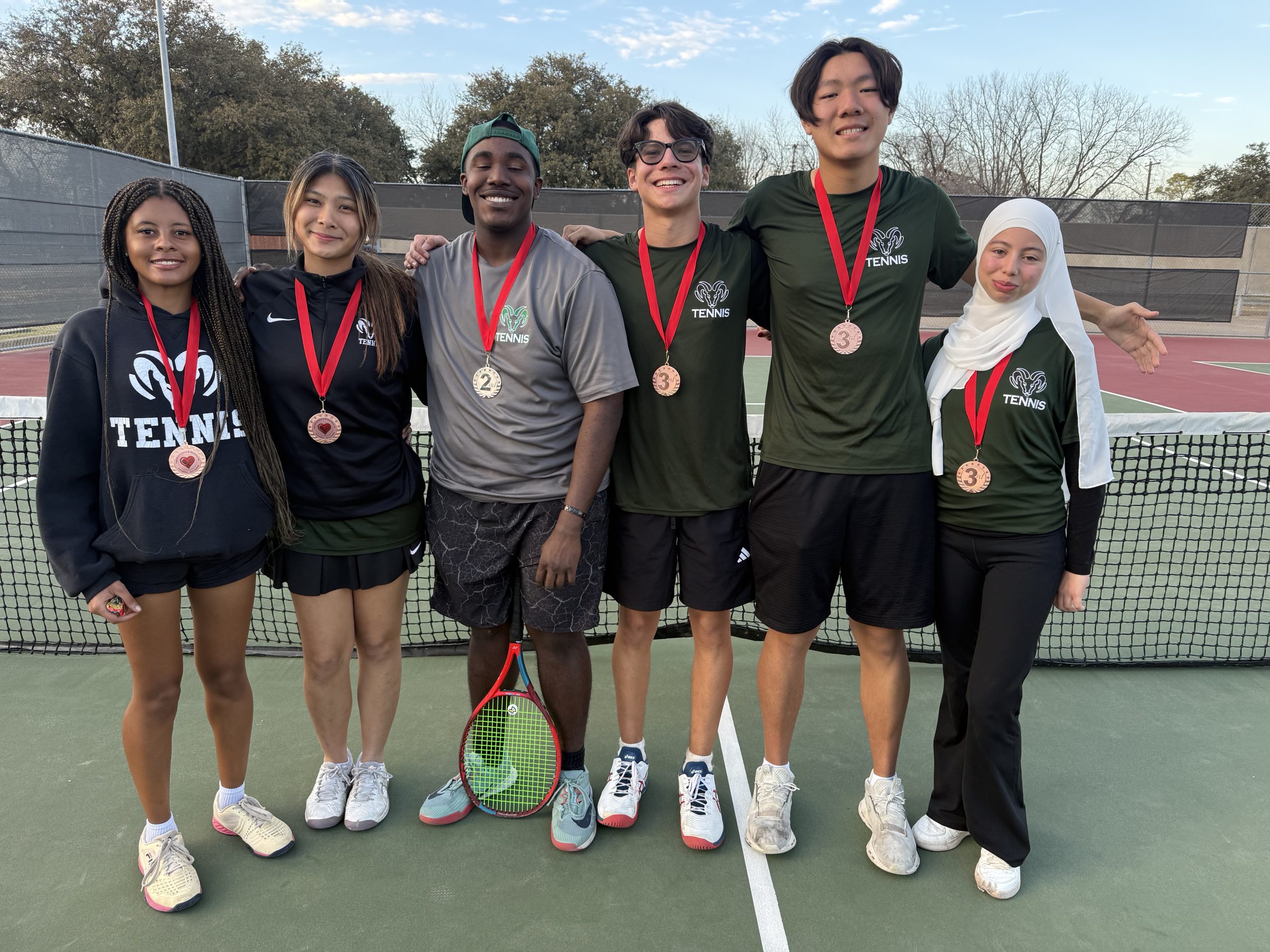 A group photo of six smiling varsity tennis players standing on an outdoor tennis court in front of the net. All six athletes are wearing medals with red ribbons around their necks. They are dressed in team gear, including dark green "TENNIS" shirts with a ram logo and black athletic wear. One player in the center holds a tennis racket.