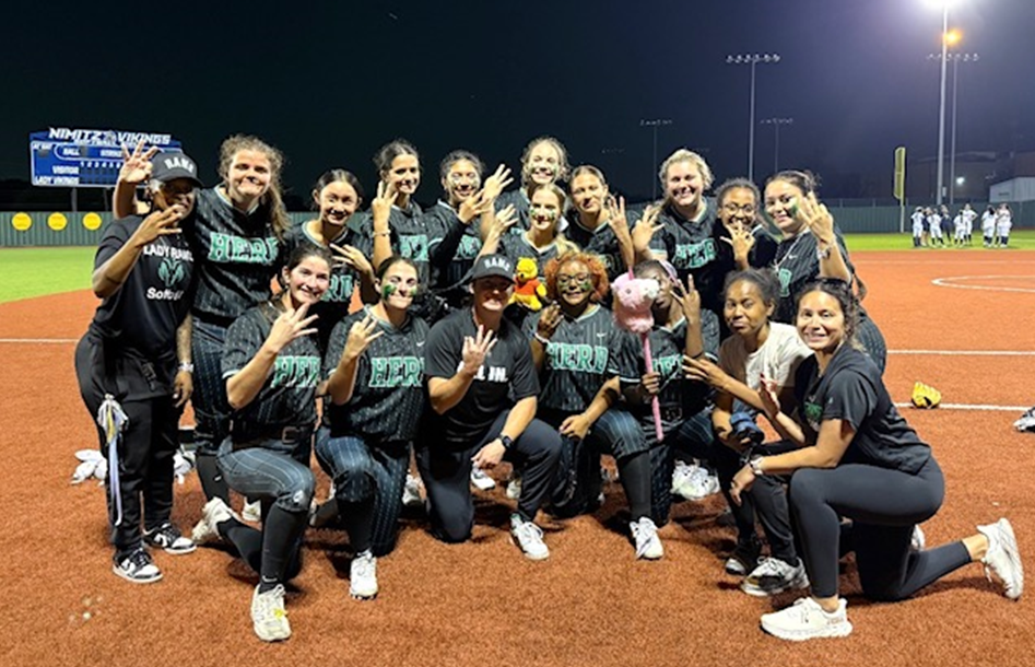 A celebratory group photo of the Lady Rams softball team and coaches on the dirt infield of a stadium at night. The players are wearing black pinstriped uniforms that say "HERD" in teal lettering, with many flashing hand signs and smiling.