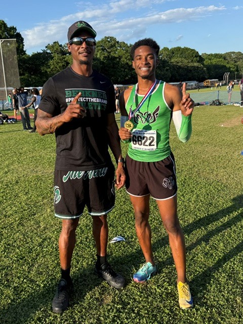 A photograph of athlete Tyler Key and his coach celebrating a win on a grassy field. Tyler, on the right, wears a green track jersey with the number 6622, black shorts, and mismatched track spikes while holding up a gold medal and pointing "number one." His coach, on the left, wears a black Berkner Athletics shirt and sunglasses, also gesturing "number one."
