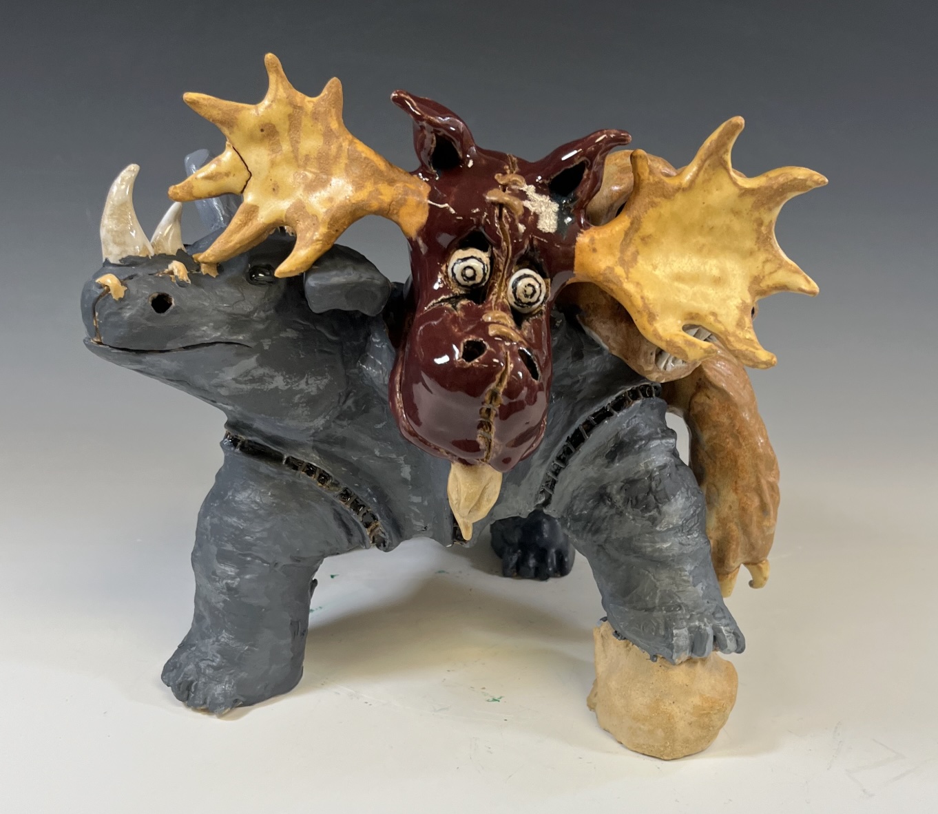 A detailed ceramic sculpture featuring a mythical creature blending traits of a rhinoceros, moose, and dragon. It features a dark grey body, yellowish antlers, white horns, and a glossy brown snout with stitched-style details and wide, round eyes.