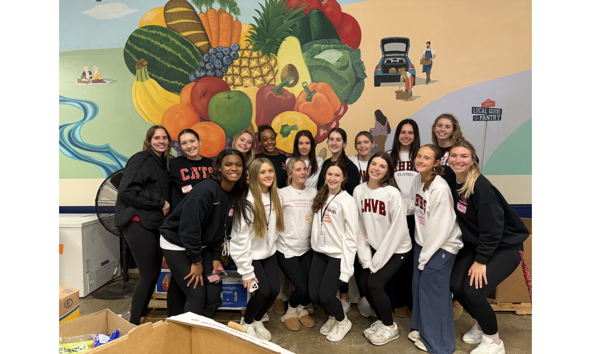 A large group of Lake Highlands High School volleyball players smiling together in front of a colorful fruit mural at a food pantry.
