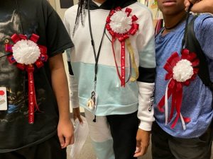 Three students showing off their DIY Homecoming mums. Modest creations of red ribbon around silk carnation with colorful ribbons streaming from the bottom of each
