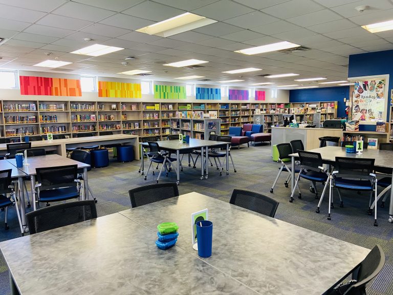 A wide shot of a school library. In the foreground are tables and chairs. In the background we see bookcases filled with books and a rainbow display on the wall above the bookcases.
