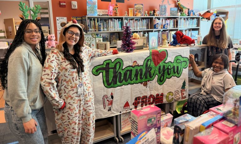 Four Pearce HS students standing behind a large "Thank You" sign and several tables overflowing with donated toys.