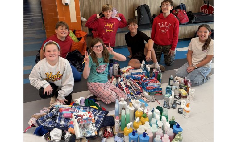 Seven elementary students sitting on the floor surrounded by a large collection of donated toiletries and clothing items.